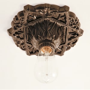 Op de afbeelding: Een zwarte metalen plafondlamp met een sierlijk ontwerp. De lamp heeft een enkele gloeilamp en een decoratieve basis.