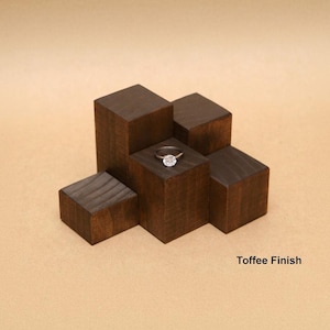 May include: A silver-toned ring with a central circular stone resting on a dark brown wooden display stand composed of five blocks of varying heights. The wood features a visible grain and is labeled with the text Toffee Finish.
