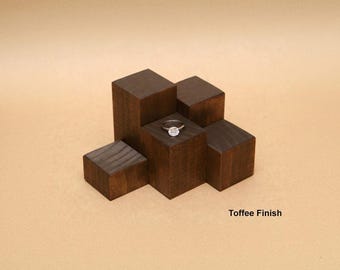 Può includere: Un anello color argento con una pietra circolare centrale poggiato su un espositore in legno marrone scuro composto da cinque blocchi di altezze variabili. Il legno presenta una venatura visibile e la scritta Toffee Finish.