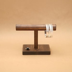 May include: A dark brown wooden T-shaped jewelry display stand with a rectangular base. A set of thin, polished silver-colored metal bangles hangs from the horizontal bar, and a single small ring with a black stone sits on the wooden base.