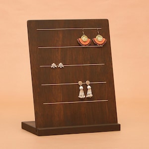 May include: A dark brown wooden earring display stand with four horizontal slots. The stand holds three pairs of earrings: orange and gold fan-shaped earrings, small silver earrings, and white beaded earrings.