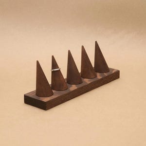 May include: A dark brown wooden ring display stand featuring five conical holders arranged in a row on a rectangular base. A single thin silver-colored metal ring is placed around the second cone from the left side of the display stand.