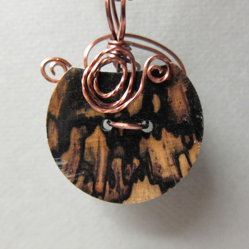 May include: A wooden button with a black and brown marbled pattern. The button is attached to a copper wire that is wrapped around the button in a decorative pattern.