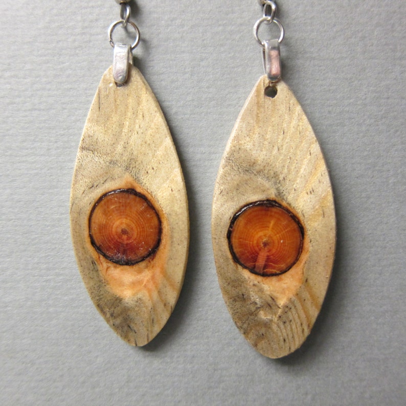 May include: A pair of wooden earrings with a natural wood finish. Each earring is shaped like a teardrop and features a circular piece of wood with a brown center and concentric rings. The earrings are attached to silver hooks.