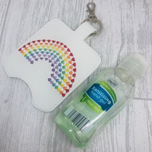 Könnte beinhalten: Weißer Kunstleder-Handdesinfektionsmittelhalter mit Regenbogenmuster, befestigt an einem silberfarbenen Schlüsselanhänger. Eine transparente Flasche Handdesinfektionsmittel-Gel mit grünem Etikett.