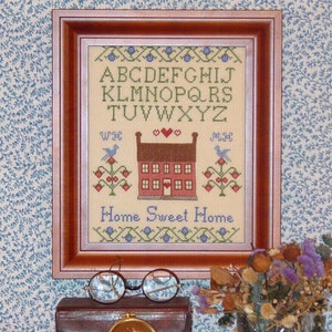 Może przedstawiać: Wzornik haftu krzyżykowego w ramie z alfabetem, domem z czerwonymi ozdobami i tekstem "Home Sweet Home". Wzornik jest wystawiony na półce z książkami i wazą z suszonymi kwiatami.