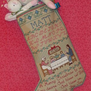 May include: A beige Christmas stocking with the name "MATT" embroidered on the front. The stocking features a cross-stitch design of a family gathered around a table with the text "God Bless This Home". The stocking is decorated with green leaves and a red bow. The stocking is filled with stuffed toys and wrapped gifts.