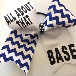 Könnte beinhalten: Weißer Cheerleader-Haar-Schleife mit blauem Glitzer-Zickzackmuster und den Wörtern "ALL ABOUT THAT" und "BASE" in Schwarz. Die Schleife wird von einem schwarzen Gummiband und einem Strassband zusammengehalten.