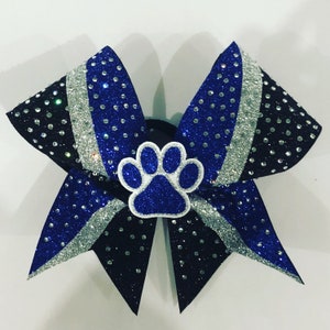 May include: A large, glittery hair bow in black, blue, and silver. The bow features a paw print in the centre and is adorned with rhinestones. The bow is designed for cheerleading or other sports.