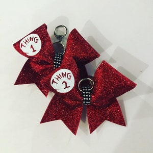 May include: Two red glitter bows with white circles that say "Thing 1" and "Thing 2" on them. The bows are attached to a silver key chain.