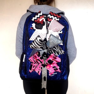 Può includere: Uno zaino blu con paillettes adornato con diversi fiocchi decorativi. I fiocchi presentano vari motivi, tra cui stampa zebrata, pois e motivi a zig-zag. Lo zaino ha una tasca laterale in rete nera e un accento glitterato argentato.