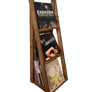 May include: A wooden record stand with three shelves. The stand is in the shape of a ladder. The top shelf has a vinyl record with the text "EMINEM CURTAIN CALL" on the cover. The middle shelf has a vinyl record with the text "AMY WINEHOUSE" on the cover. The bottom shelf has a vinyl record with a colorful cover.