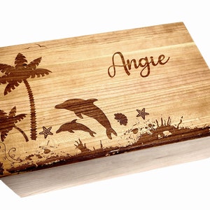 May include: A wooden box with a personalized inscription "Angie" and a laser-engraved design of palm trees, dolphins, and seashells. The box has a natural wood finish.