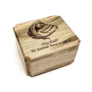 May include: A wooden box with a baseball glove engraving on the lid. The text "Hey Dad? Ya wanna have a catch?" is etched below the glove. The box is light brown with a natural wood grain.
