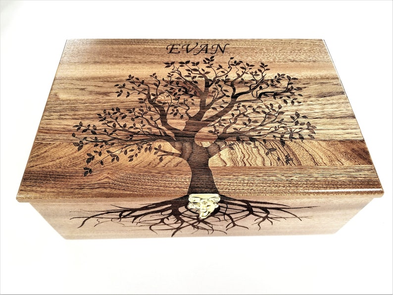 May include: A wooden box with a tree of life design engraved on the lid. The tree has a thick trunk and many branches with leaves. The box has a brass latch and the name "EVAN" is engraved on the lid.