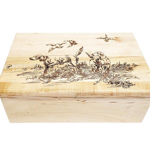 Puede incluir: Una caja de madera con tapa con bisagras. La tapa está decorada con una imagen pirograbada de dos perros y tres pájaros en un campo.