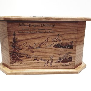 May include: A wooden cremation urn with a laser engraved image of a mountain scene with a deer in the foreground. The urn is inscribed with the name "Jeffery Eugene Defibaugh" and the dates "December 51, 1964 - September 15, 2020". The inscription also reads "Our Loving Husband, Father, And Brother".
