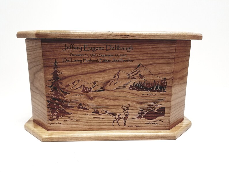 May include: A wooden cremation urn with a laser engraved image of a mountain scene with a deer in the foreground. The urn is inscribed with the name "Jeffery Eugene Defibaugh" and the dates "December 51, 1964 - September 15, 2020". The inscription also reads "Our Loving Husband, Father, And Brother".