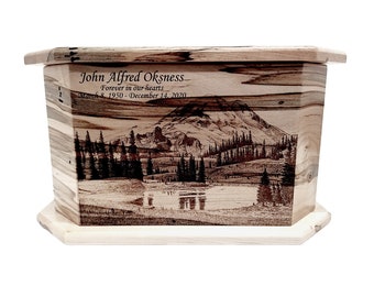 May include: A wooden cremation urn with a woodburned image of a mountain lake and forest scene. The urn has a lid and is engraved with the text "John Alfred Oksness Forever in our hearts March 8, 1950 - December 14, 2020".