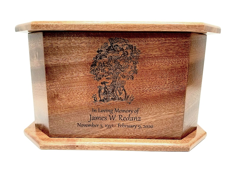 May include: A wooden cremation urn with a tree of life design engraved on the front. The urn is inscribed with "In Loving Memory of James W. Redanz November 3, 1952 - February 9, 2020".