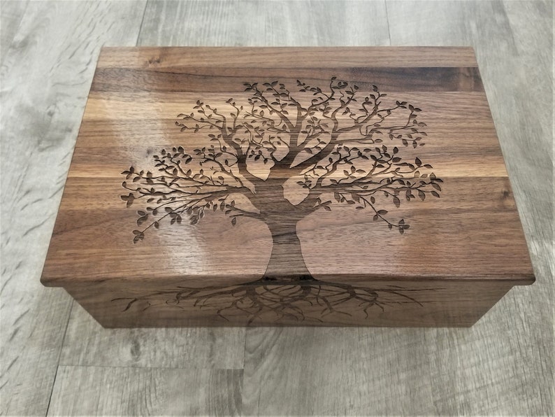 May include: A wooden box with a tree design carved into the lid. The tree has a thick trunk and many branches with leaves. The roots of the tree are also carved into the wood.