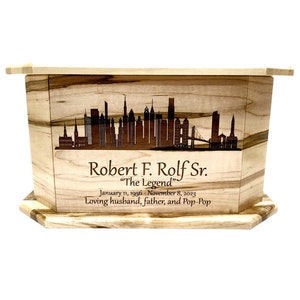 May include: A wooden cremation urn with a cityscape design and the inscription "Robert F. Rolf Sr. "The Legend" January 11, 1956 - November 8, 2023 Loving husband, father, and Pop-Pop"