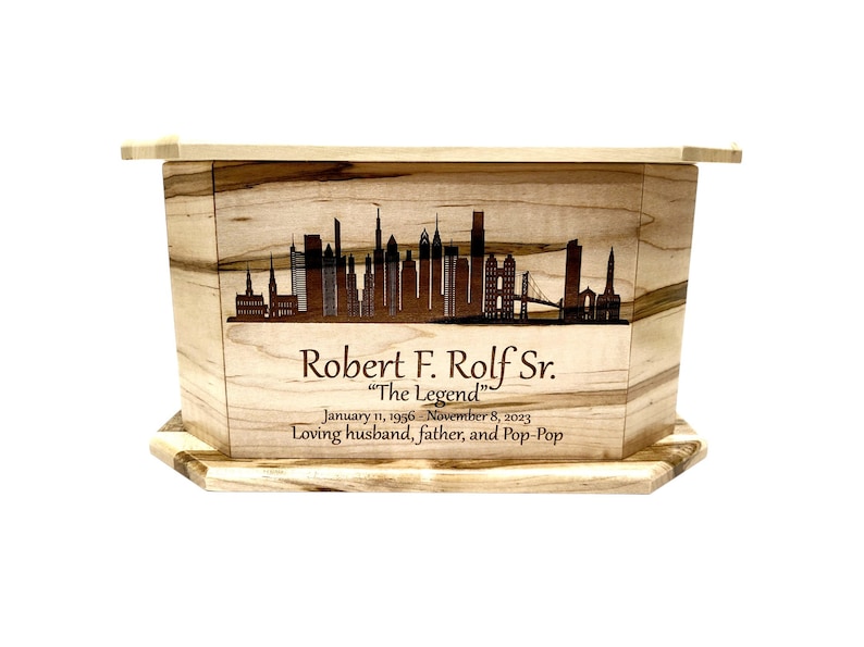 May include: A wooden cremation urn with a cityscape design and the inscription "Robert F. Rolf Sr. "The Legend" January 11, 1956 - November 8, 2023 Loving husband, father, and Pop-Pop"
