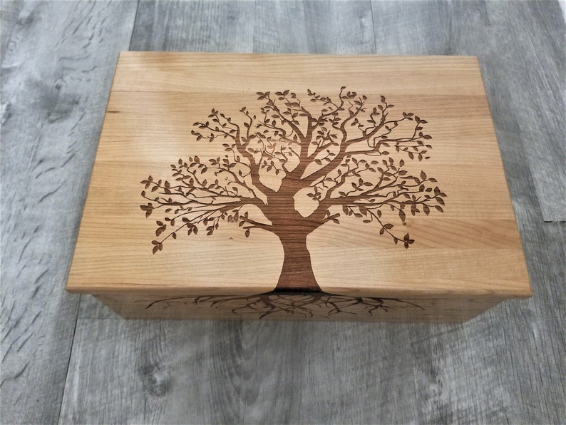 May include: A wooden box with a tree design carved into the lid. The tree has a dark brown trunk and branches with leaves. The box is made of light wood and has a natural finish.