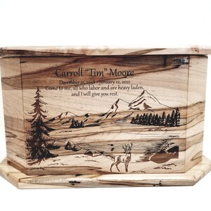 May include: A wooden cremation urn with a laser engraved image of a mountain scene with a deer in the foreground. The urn is inscribed with the name "Carroll "Tim" Moore", dates "December 21, 1998 January 12, 2021", and the quote "Come to me, all who labor and are heavy laden, and I will give you rest."