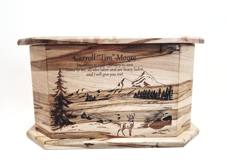 May include: A wooden cremation urn with a laser engraved image of a mountain scene with a deer in the foreground. The urn is inscribed with the name "Carroll "Tim" Moore", dates "December 21, 1998 January 12, 2021", and the quote "Come to me, all who labor and are heavy laden, and I will give you rest."