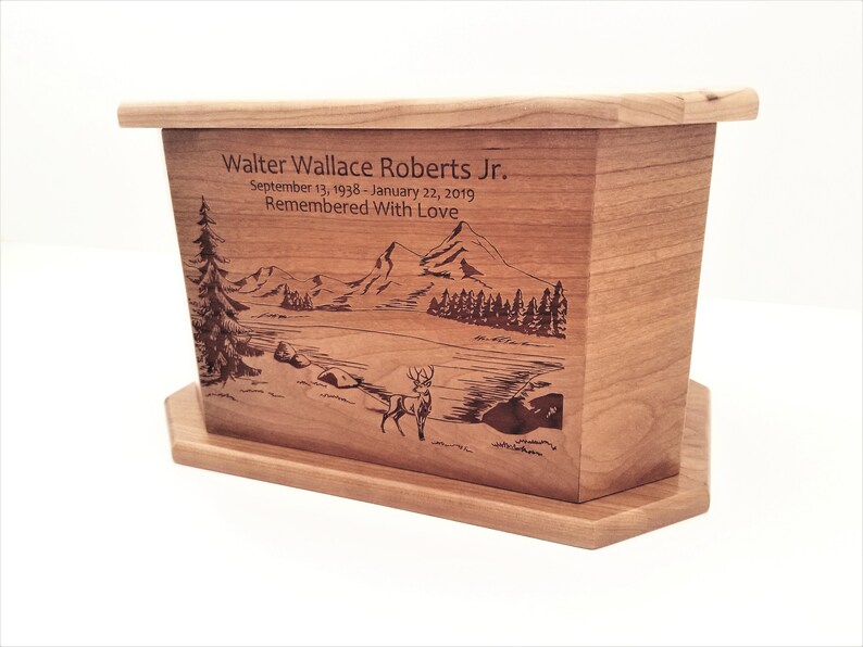 May include: A wooden cremation urn with a laser engraved image of a mountain scene with a deer in the foreground. The urn is inscribed with the name "Walter Wallace Roberts Jr." and the dates "September 13, 1938 - January 22, 2019". The inscription also includes the phrase "Remembered With Love".