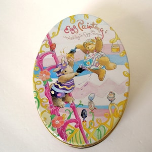 May include: A round, oval-shaped tin with a colorful illustration of two animals painting eggs. The tin has the text "Egg Painting" and "Walking in Egg Shells" on it.