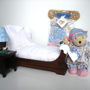 May include: A miniature bedroom set featuring a wooden bed with white bedding, a nightstand with a blue lampshade, and a teddy bear wearing pajamas and a hat. A package of matching bedding is also visible.