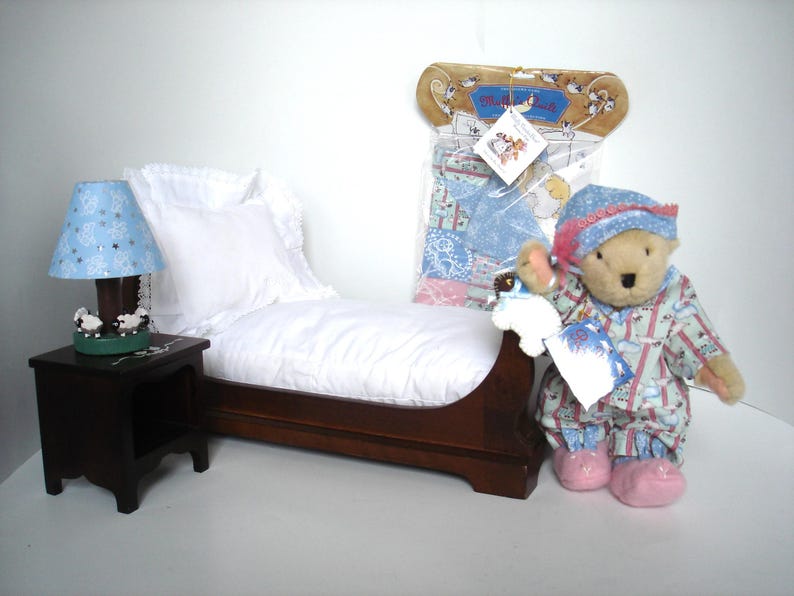 May include: A miniature bedroom set featuring a wooden bed with white bedding, a nightstand with a blue lampshade, and a teddy bear wearing pajamas and a hat. A package of matching bedding is also visible.
