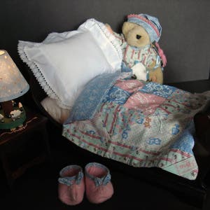 May include: A miniature bedroom scene featuring a teddy bear in pajamas and a nightcap, nestled under a patchwork quilt. A white pillow with lace trim rests against the headboard. A small lamp with a sheep design and pink booties complete the cozy setting.