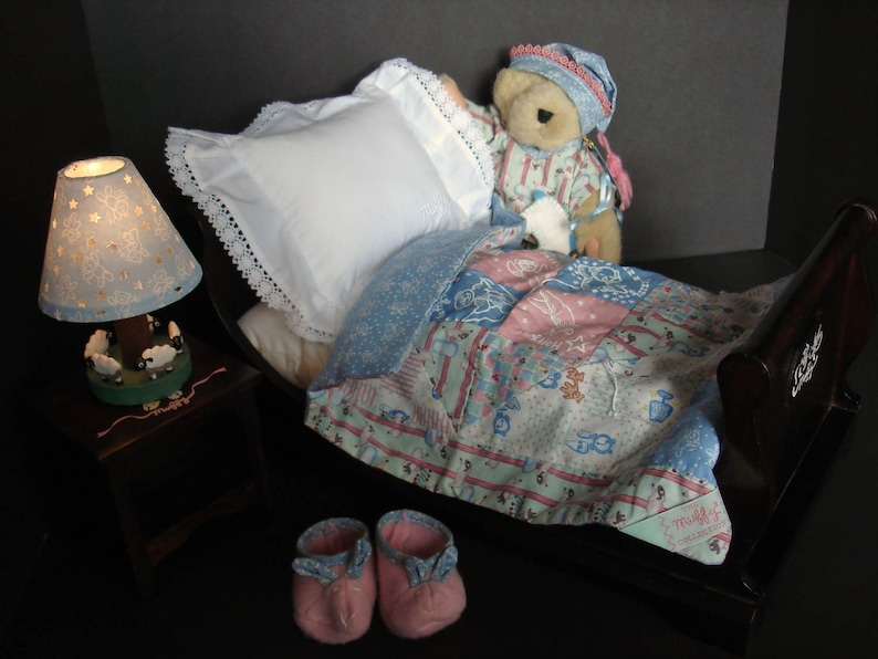 May include: A miniature bedroom scene featuring a teddy bear in pajamas and a nightcap, nestled under a patchwork quilt. A white pillow with lace trim rests against the headboard. A small lamp with a sheep design and pink booties complete the cozy setting.