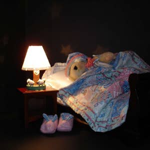 May include: A cozy bedroom scene with a teddy bear in bed under a patterned quilt. A lit lamp with a heart design sits on a small table next to the bed. Pink slippers are on the floor.