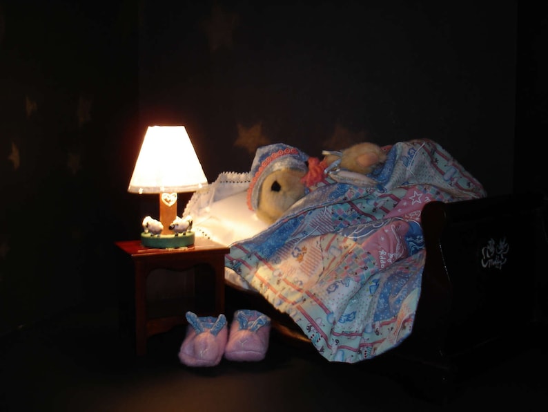 May include: A cozy bedroom scene with a teddy bear in bed under a patterned quilt. A lit lamp with a heart design sits on a small table next to the bed. Pink slippers are on the floor.