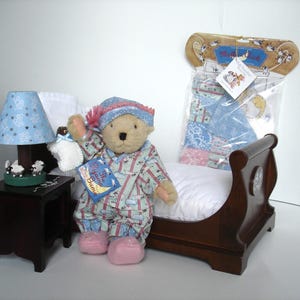 May include: A teddy bear wearing pajamas and a nightcap, holding a bottle and a book titled "The Pajama Game." The bear is sitting on a wooden bed with a white pillow. A small lamp with a blue shade and a nightstand are also present.