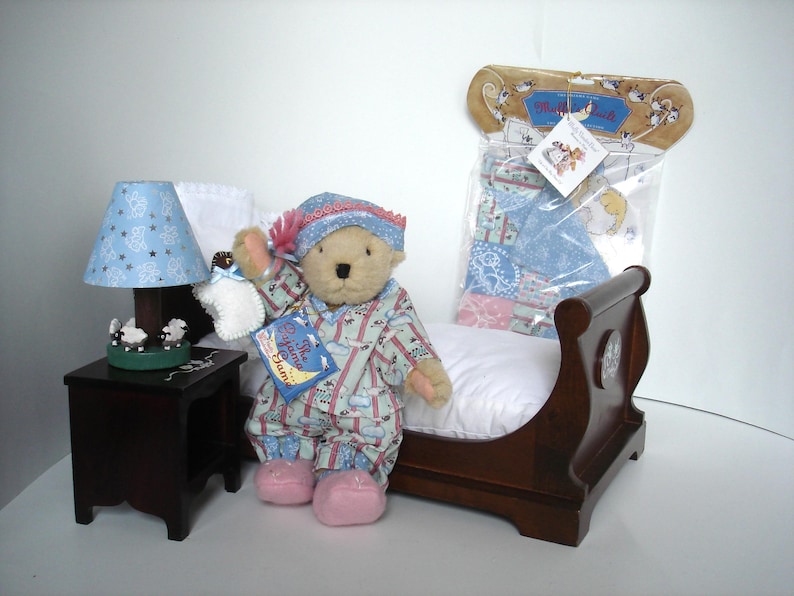 May include: A teddy bear wearing pajamas and a nightcap, holding a bottle and a book titled "The Pajama Game." The bear is sitting on a wooden bed with a white pillow. A small lamp with a blue shade and a nightstand are also present.