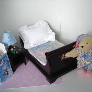 May include: A miniature bedroom set with a dark brown bed, white pillow, and patterned bedding. A blue lamp with star cutouts sits on a black nightstand. A teddy bear in pajamas and a box with a moon and stars design complete the scene.