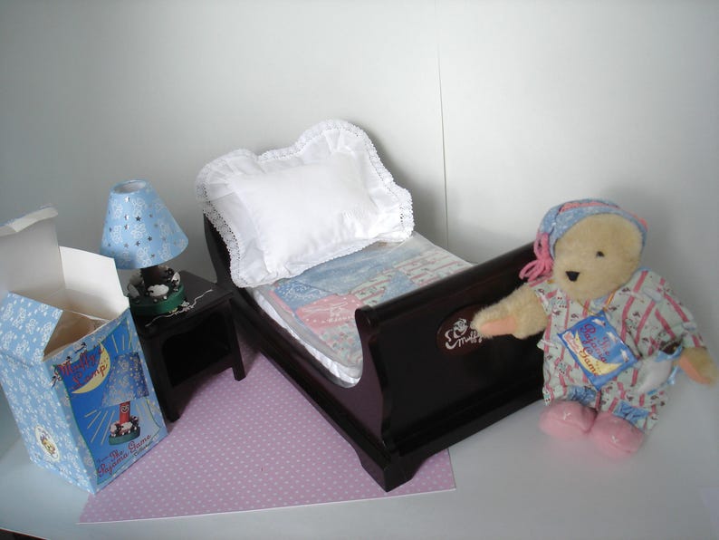 May include: A miniature bedroom set with a dark brown bed, white pillow, and patterned bedding. A blue lamp with star cutouts sits on a black nightstand. A teddy bear in pajamas and a box with a moon and stars design complete the scene.
