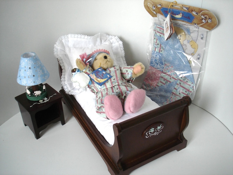 May include: A miniature bedroom set featuring a teddy bear in pajamas lying in a wooden bed with a white pillow and blanket. A blue lamp with star cutouts sits on a nightstand. A packaged quilt is also present.