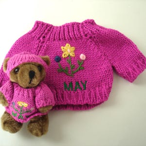 May include: A brown plush teddy bear wearing a matching magenta knitted sweater and hat, alongside a larger magenta knitted sweater. Both sweaters feature embroidered green stems, a yellow flower, and the word MAY stitched in green thread.