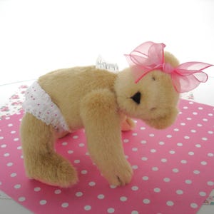 May include: A plush, tan-colored teddy bear wearing a white diaper with red polka dots and a sheer pink bow. The bear is posed on a pink polka dot background, suggesting a baby or nursery theme.