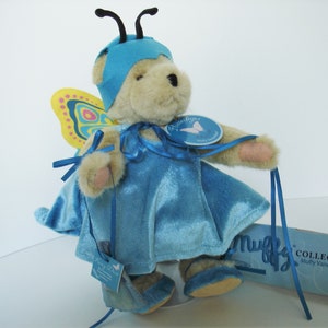 May include: A blue and yellow butterfly costume for a plush bear. The bear is wearing a blue dress and a blue butterfly hat. The bear is holding a blue ribbon in its paw. The bear is sitting on a white surface. The bear is part of the Muffy VanderBear collection.