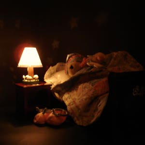 May include: A miniature bedroom scene with a lit lamp on a small table, a doll under a blanket, and a pair of orange shoes. The lamp has a white lampshade and a wooden base. The background is dark with star-shaped patterns.