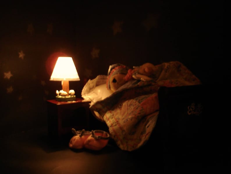 May include: A miniature bedroom scene with a lit lamp on a small table, a doll under a blanket, and a pair of orange shoes. The lamp has a white lampshade and a wooden base. The background is dark with star-shaped patterns.