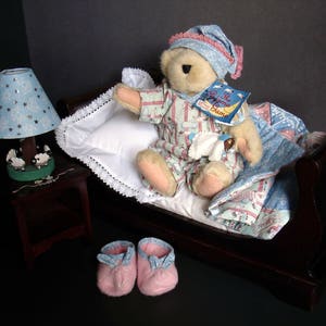 May include: A teddy bear wearing pajamas and a nightcap sits in a miniature bed with a pillow and blanket. A bedside lamp with a blue shade and a pair of pink slippers complete the scene. The bear holds a book titled "The Dream Game".