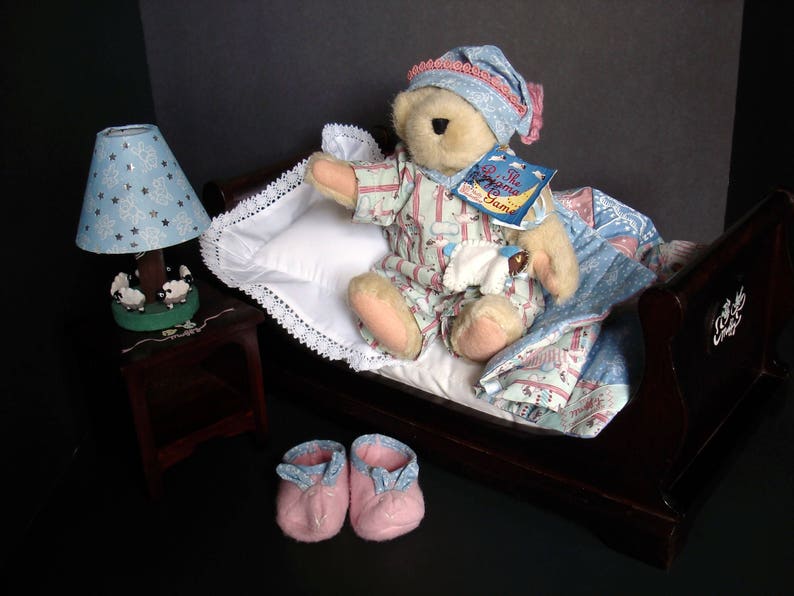 May include: A teddy bear wearing pajamas and a nightcap sits in a miniature bed with a pillow and blanket. A bedside lamp with a blue shade and a pair of pink slippers complete the scene. The bear holds a book titled "The Dream Game".